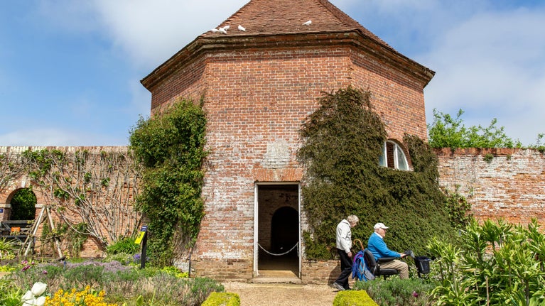 Visitors explore the Walled Garden at Felbrigg, passing a brick garden building covered in climbing plants. Colourful flower borders and neatly trimmed hedges line the path in the foreground.